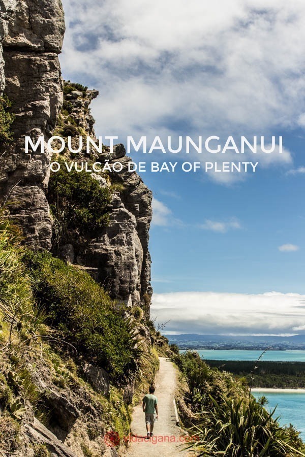 Um homem sobe os entornos do Mount Maunganui, um vulcão extinto na Ilha Norte da Nova Zelândia. Uma parede imensa de pedras cerca a trilha de um lado. A trilha dura 1 hora ida. Lá embaixo, uma praia azul. O céu está azul, com algumas nuvens baixas. 