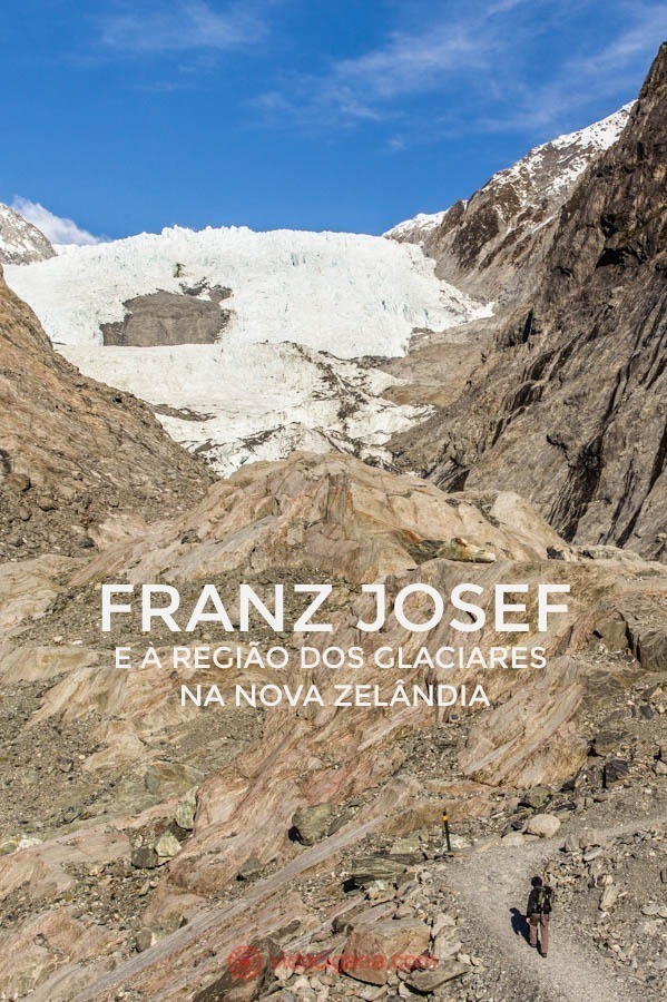 A trilha do glaciar Franz Josef, o mais famoso da Nova Zelândia, fica na Ilha Sul do país. Leva uma hora a ida até o mirante. Um homem caminha pela trilha em meio a rochas cor de terra até o mirante, mas o glaciar está lá no fundo, tímido, mas branco, entre duas montanhas. O céu está azul.