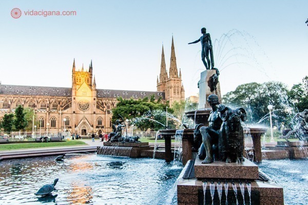 O que fazer em Sydney: Uma catedral iluminada pelo por do sol no fundo da imagem com uma estátua dentro de uma fonte em primeiro plano