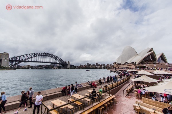 O que fazer em Sydney: O Circular Quay, cheio de mesas na beira do mar, com a Ópera de Sydney do lado direito da foto e a Harbour Bridge, a ponte do porto, do lado esquerdo