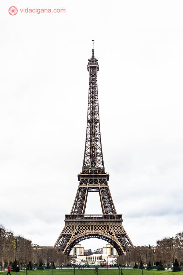 A Torre Eiffel em Paris na frente do Campo de Marte com grama verde. O céu na foto está nublado e branco, com um clima bastante frio. As árvores estão secas e sem folhas, típicas do inverno, com uma tonalidade marrom.