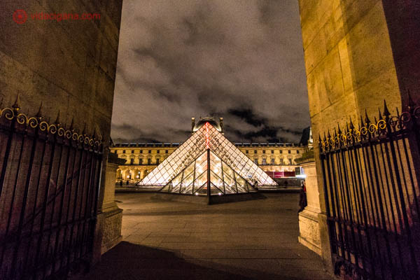 Pirâmide do Museu do Louvre de vidro com raio laser vermelho em seu interior emoldurada por duas paredes com portões. Foto tirada a noite, com o cpeu nublado, cheio de nuvens passando.