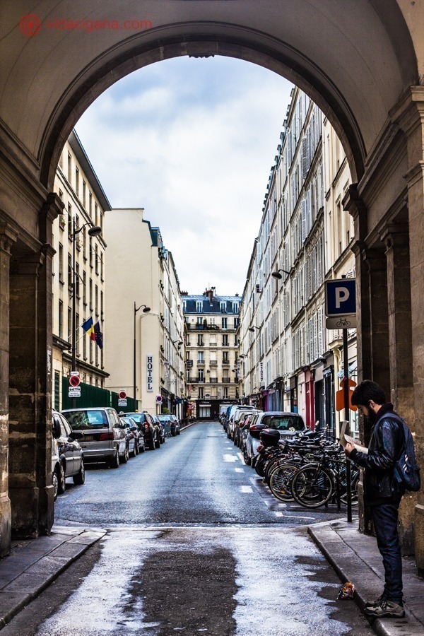 Um rua em Paris, onde é possível ver os apartamentos através de um arco na entrada da rua. Na frente do arco, um homem vestindo casaco preto e calça jeans e carregando uma mochila azul, está lendo um livro. Vários carros estão estacionados na calçada. O céu está azul com nuvens brancas. O clima é frio.