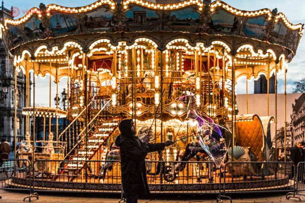 Carrossel dourado e clássico em Paris, com morador de rua fazendo bolhas de sabão na frente dele. O homem veste um casaco preto com capuz. O carrossel é no estilo antigo, com cavalos e luzes com lâmpadas bolinhas.