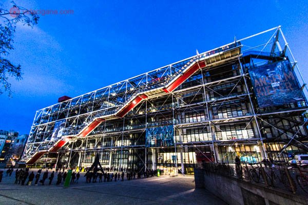 O exterior do Centro Pompidou, em Paris, com sua fachada moderna, feita de ferro e com uma escadaria vermelha. O céu está azul e está de noite. Tudo iluminado e uma fila é vista na frente do museu.