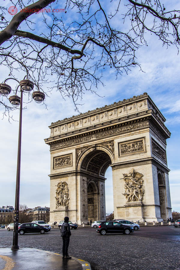 O Arco do Triunfo em Paris num dia de sol. A foto está emoldurada com galhos secos e sem folhas de uma árvore. Também vemos um poste com quatro lâmpadas e um homem em frente ao Arco, do outro lado da calçada.