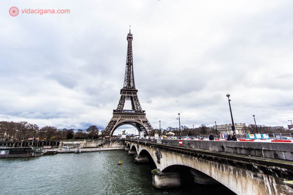 A Torre Eiffel vista ao fundo. Na frente encontra-se uma ponte em Paris e o rio Sena. O céu está nublado.
