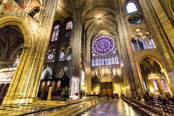 O interior da Catedral de Notre Dame de Paris, na França. Seu interior é enorme, no estilo gótico, cheio de vitrais coloridos e uma rosetta em seu alto que ilumina o interior da igreja com uma luz lilás. 