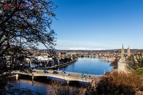 A cidade de Inverness, na Escócia, vista de cima. Um lindo rio azul corre, com uma ponte sobre ele. Carros passam por lá. Prédios baixos estão dos dois lados do rio. Uma árvore se encontra do lado esquerdo da foto. O céu está azul.