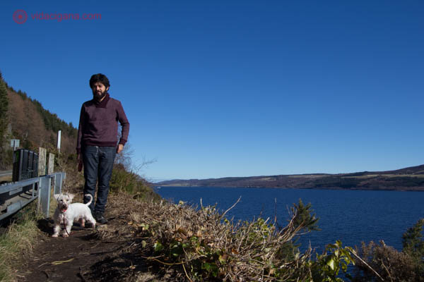 Um homem com um schnauzer branco na beira do Lago Ness, na Escócia. O lago é azul profundo, o céu também. Montanhas estão no fundo.