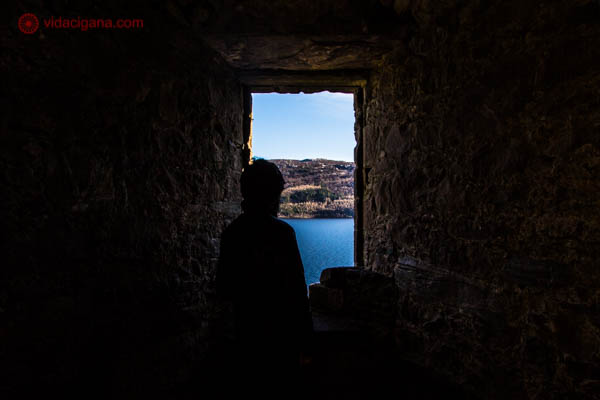 Homem se encontra numa janela do castelo de urquhart, na Escócia, observando o Lago Ness ao fundo. Só é possível ver a silhueta. O lago é azul e o céu também. 