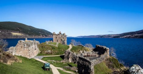 O Castelo de Urquhart, na Escócia, com o Lago Ness, ou Loch Ness, no fundo. O céu está azul sem nenhuma nuvem. O lago possui uma cor azul profunda, lindíssima. Montanhas estão dos dois lados do lago. As ruínas do castelo se encontram em primeiro plano, feito de pedras, muros e torres. Algumas árvores estão secas no local. O castelo fica numa área gramada e verde.