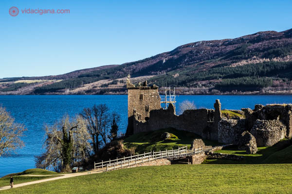 O Castelo de Urquhart em primeiro plano, em ruínas, na Escócia. Ele é feito de pedras. Uma ponte liga o castelo ao outro lado. Árvores secas estão no local. Atrás do castelo está o Lago Ness, de cor azul. Ao fundo, montanhas. O céu está azul.
