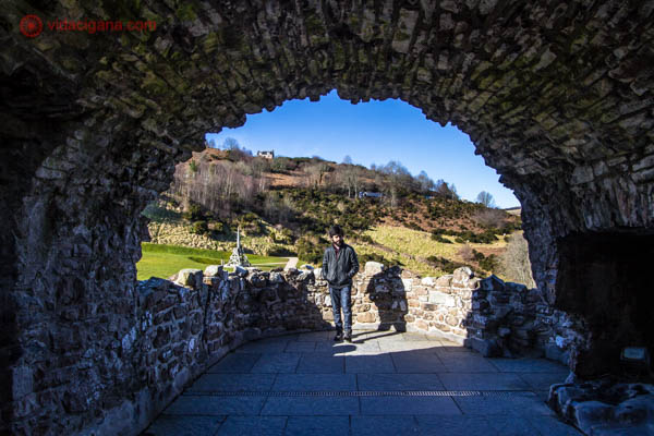 Um homem moreno está parado numa sacada do castelo de Urquhart, na Escócia. A foto é emoldurada por um arco de pedras. Ao fundo, uma montanha cheia de árvores secas e grama se encontra. O céu é azul.