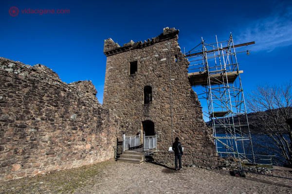 Grant Tower, a maior torre do castelo de urquhart, na Escócia. Um homem está parado na frente da construção, que é uma ruína com andaimes do lado direito. O céu está azul, e ao fundo, o Lago Ness.