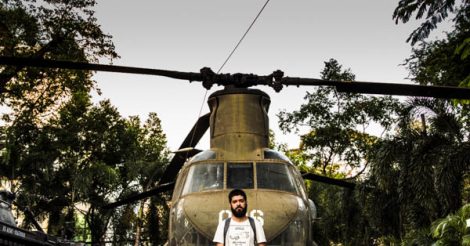 Homem moreno de barba, vestindo uma blusa branca e carregando uma mochila, posa na frente de um helicóptero americano capturado pelos vietnamitas durante a Guerra do Vietnã. Essa foto foi tirada na Cidade de Ho Chi Minh, antiga Saigon, capital do Vietnã do Sul na época. O helicóptero faz parte da coleção de antigos equipamentos de guerra do War Remnants Museum.
