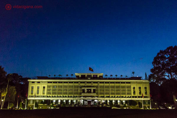 Na foto vemos o Palácio da Reunificação, prédio histórico importante que fica na Cidade de Ho Chi Minh. Foi nesse prédio que a Guerra do Vietnã acabou. O Palácio tem formato retangular, no estilo arquitetônico moderno, com uma bandeira do Vietnã no topo. A foto foi tirada durante a noite, com o céu limpo e azulado. O prédio está iluminado com uma cor amarela.
