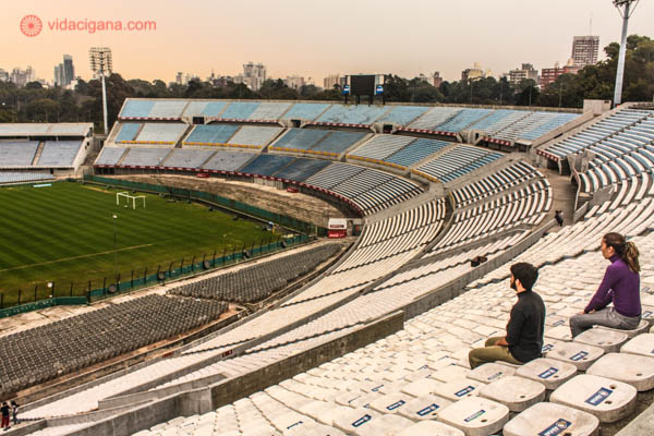 Um homem e uma mulher sentados em fila no Estádio Centenário, em Montevidéu, no Uruguai. O estádio é imenso e possui vários cores em suas arquibancadas. O céu está nublado, mas possui cores do pôr do sol, em tom alaranjado. O campo de futebol é verde e pode ser visto lá embaixo.