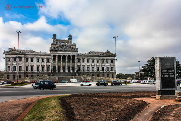 O Palácio Legislativo de Montevidéu. Ele tem um estilo clássico, com sua fachada repleta de colunas e frontões. O Palácio se encontra ao fundo. Na frente, uma praça com um pouco de grama verde e chão de terra batida fica em primeiro plano. O céu está parcialmente nublado, com alguns trechos azuis.
