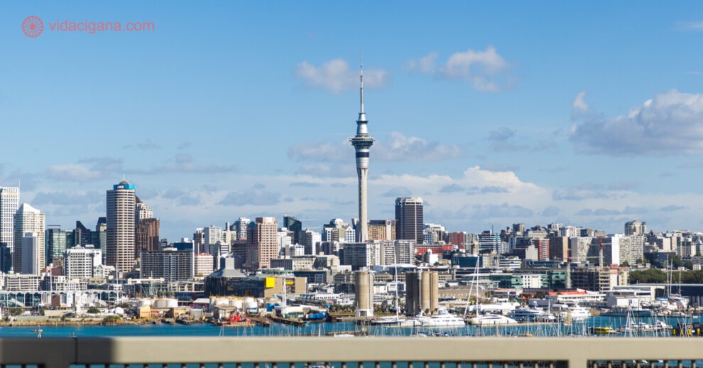 A cidade de Auckland vista do outro lado da baía, com todos os seus prédios, com destaque ao Sky Tower, a torre mais alta do país. O céu está azul, com algumas poucas nuvens brancas. A cidade está na beira da água.