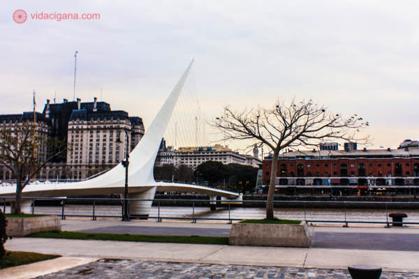 A Ponte de la Mujer em Puerto Madero, em Buenos Aires. A ponte está do lado esquerdo e possui uma forma bastante diferente. Ela é branca e estaiada, com vários cabos em sua extensão. Uma árvore está do lado direito da foto. O céu está nublado. Prédios aparecem ao fundo.