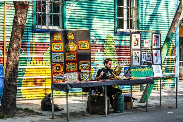 Artista de rua no Caminito, em Buenos Aires. Ele está pintando sentado em uma cadeira, com mesa no meio da rua. Várias placas coloridas enfeitam sua mesa. Atrás, a parede de casas de todas as cores. 