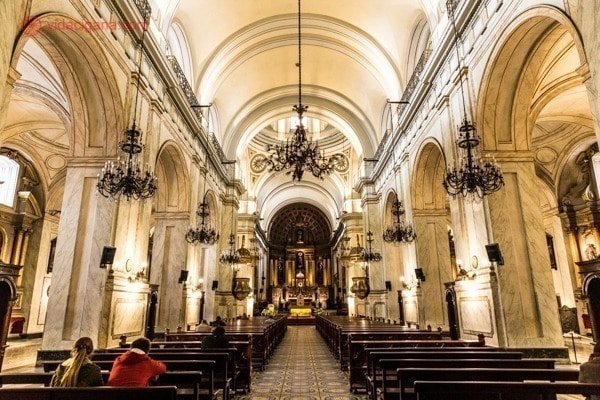 O interior da Catedral Metropolitana de Montevidéu. Muito bonita, com paredes brancas, mas iluminação amarelada, dando um ar acolhedor. Vários bancos com poucas pessoas sentadas, vários candelabros negros pendurados.