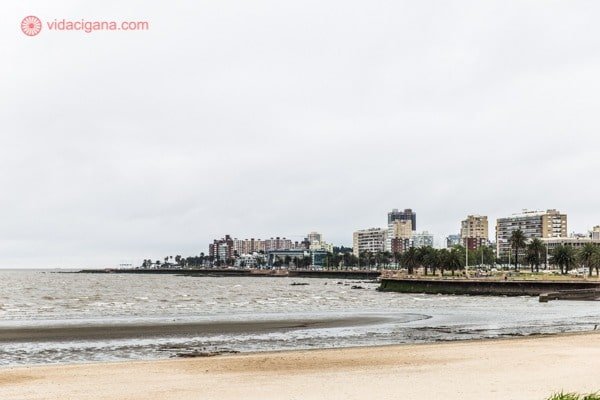 Praia em Montevidéu, no Uruguai, na beira do Rio de la Plata. Ao fundo se vê a cidade, com seus prédios. O mar tem tom acizentado e a areia é amarelada. O céu está nublado.