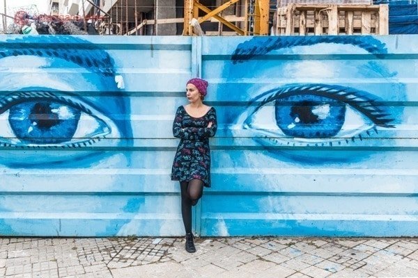 Mulher de toca rosa e vestido preto estampado, usando meia calça preta e tênis preto está apoiada em um mural de graffiti em Montevidéu, no Uruguai. Na arte podemos ver dois olhos abertos. O mural é todo azul.