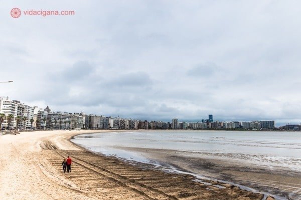 Uma praia em Montevidéu, no Uruguai, com uma grande faixa de areia e o rio de la Plata com uma tonalidade escura. O céu está nublado e ao fundo podem ser vistos os prédios na Rambla.