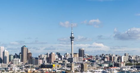 A cidade de Auckland vista do outro lado da baía, com todos os seus prédios, com destaque ao Sky Tower, a torre mais alta do país. O céu está azul, com algumas poucas nuvens brancas. A cidade está na beira da água.