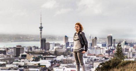 Mulher ruiva em cima da borda do vulcão Mount Eden, em Auckland, na Nova Zelândia, com vista para a cidade lá ao fundo, com a Sky Tower, uma torre pontiaguda, se destacando lá ao longe. O céu está nublado. A cor predominante na foto é o cinza.