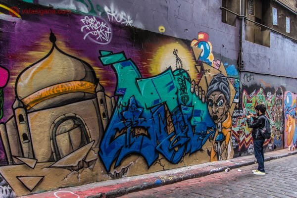 Um homem de preto tirando fotos de uma parede com grafites em Melbourne, na Austrália. O mural é bem colorido, cobrindo quase a parede toda.
