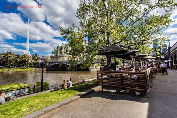 Onde ficar em Melbourne: A região de Southbank, em Melbourne, num dia ensolarado, com pessoas sentadas na grama vendo o rio, vários cafés na beira do rio, muitas árvores, a ponte ao fundo, e um prédio com um estilo bastante peculiar. 