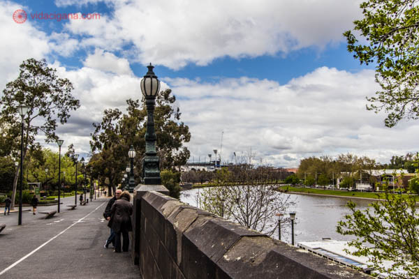 Onde ficar em Melbourne: O rio Yarra, em Melbourne, com pessoas conversando encostadas na mureta na beira do rio. O céu está azul com nuvens brancas.