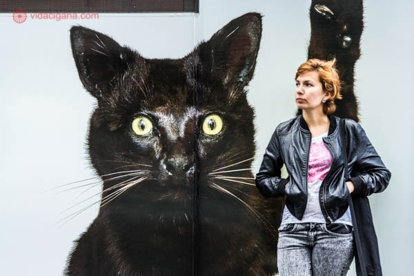 Mulher ruiva com jaqueta de couro encostada em um mural com a foto de um gato preto gigante, em Melbourne, na Austrália.