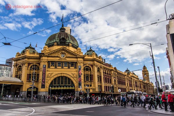 O prédio do Flinders Street Railway Station, em Melbourne, na Austrália, com sua cor amarela e sua arquitetura antiga. Com sua cúpula verde e estilo ligeiramente indiano, o prédio fica numa área bem movimentada, onde várias pessoas cruzam as faixas de trânsito bem em frente. O céue stá azul com algumas nuvens brancas e algumas cinzas.