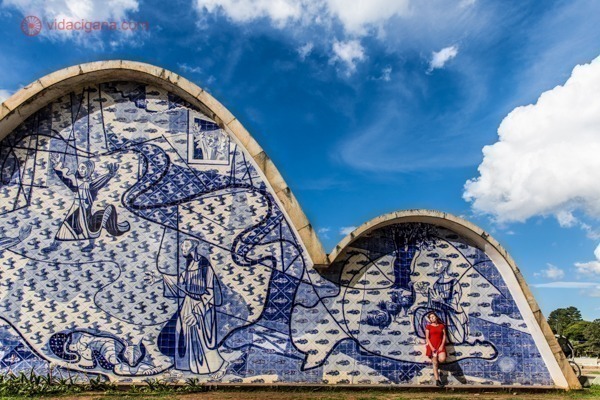 A igreja da Pampulha com seus enormes painéis produzidos por Cândido Portinari. Todos em tons de azul e branco. Uma mulher vestida de vermelho se destaca na foto e está encostada no mural. O céu é azul com algumas nuvens.