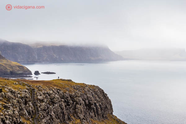 Um homem na beira do penhasco em Neist Point, na Ilha de Skye, na Escócia. Lá atrás, montanhas cobertas de neblina dão um ar melancólico ao lugar.