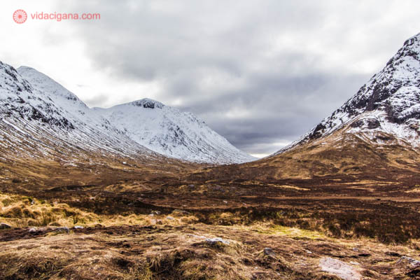 Montanhas nevadas nas Highlands, as Terras Altas da Escócia, no Reino Unido. O clima da foto é melancólico, com o céu nublado. As montanhas são altas, cor de terra, com uma vegetação rasteira e seca. O topo das montanhas estão cheios de neve.