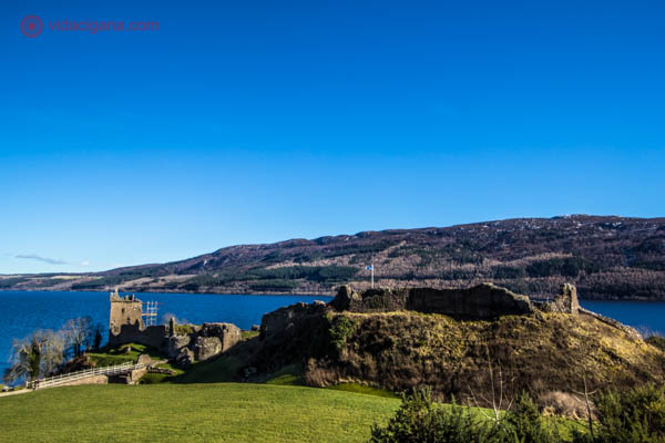 O Castelo de Urquhart a frente do Lago Ness, nos Highlands escoceses. O céu está azul, o lago também.