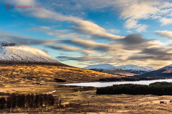 Montanhas nevadas nos Highlands, as Terras Altas da Escócia. O dia está ensolarado, com uma luz amarelada e tímida. O céu está azul, com algumas nuvens. Um lago se encontra na frente das montanhas.