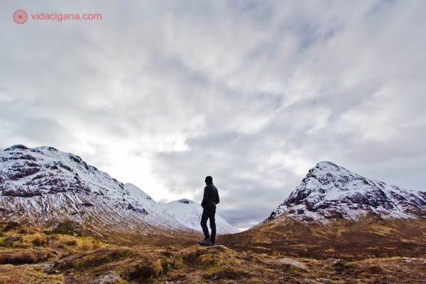 A silhueta de um homem entre duas montanhas nevadas nos Highlands, as Terras Altas da Escócia. O céu está nublado e as montanhas cheias de neve no seu topo.