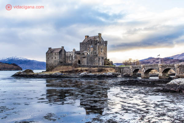 O Castelo de Eilean Donan, nos Highlands, as Terras Altas da Escócia. O castelo fica em uma ilhota cercada de um lago. Uma ponte leva da terra até a área do castelo. Ele é feito de pedra, assim como sua ponte. O céu está nublado, com alguns pontos azuis. Montanhas estão ao fundo.