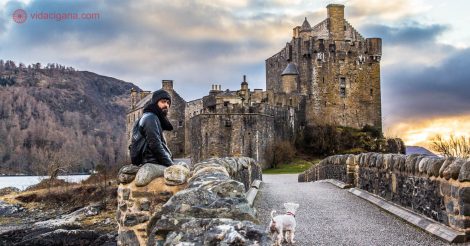 Homem vestindo jaqueta preto e gorro descansa sobre mureta de pedra enquanto segura pela correia a coleira de um cachorro branco da raça scnauzer. A cena ocorre em frente ao Castelo de Eilean Donan, próximo à ilha de skye nas Highlands da Escócia.