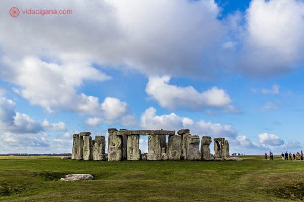 O monumento de Stonehenge, na Inglaterra, feito de blocos de pedra imensos formando um círculo em um gramado verde. O céu é azul com muitas nuvens.