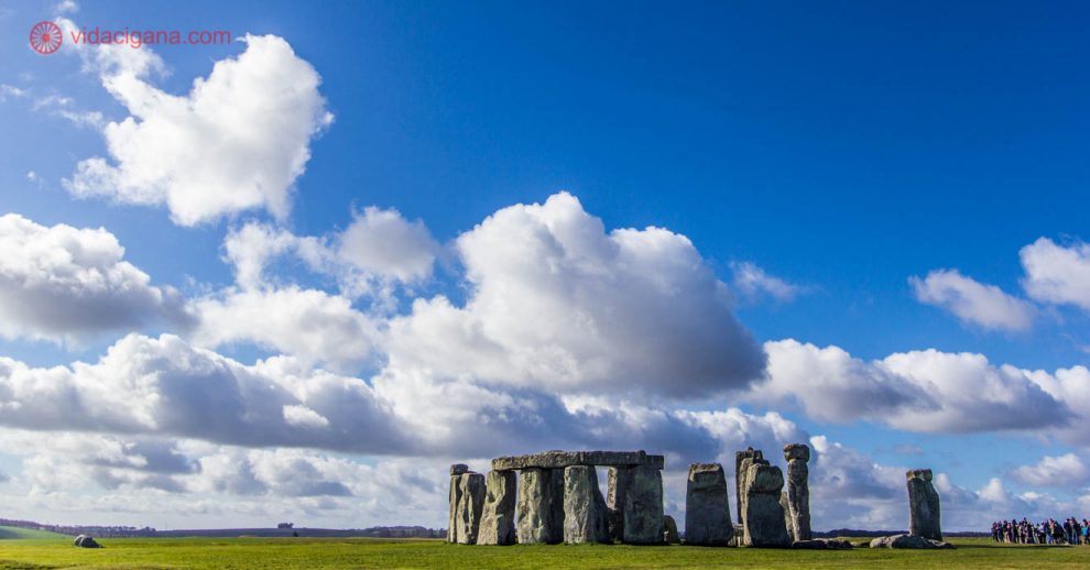 O monumento de Stonehenge, na Inglaterra, feito de blocos de pedra do período neolítico. O círculo de pedra se encontra em um campo de grama verde imenso. O céu está muito azul, com nuvens brancas que voam pro lado esquerdo da foto.