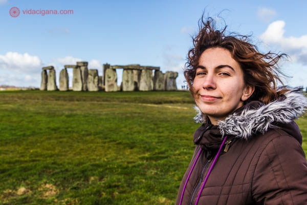 Uma mulher morena na frente do monumento de Stonehenge, na Inglaterra. Ela está sorrindo, usando uma roupa de inverno, com os cabelos ao vento. O céu está azul, a grama é verde, e o monumento está lá atrás.