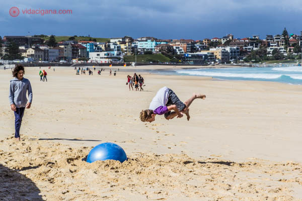 Crianças brincam de dar piruetas no ar em cima de uma bola em Bondi Beach. Elas brincam na areia, na frente do mar. Ao fundo, mansões na encosta da praia.