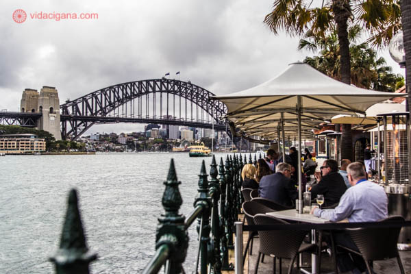 Várias pessoas comem em mesas na beira do Circular Quay, em Sydney. De lá é possível ver a Sydney Bridge do lado esquerdo da foto. As pessoas comem em mesas com guarda sol, com aquecedores do lado, na frente de uma grade. O dia está nublado.